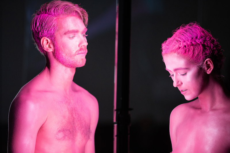 A dancers, their bodies and faces cover in hot pink clay, stand near one another. One looks down while their other look directly at his partner.